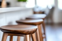 Modern wooden bar stools with smooth curved seats lined up at kitchen counter, selective focus on foreground stool creates depth in bright interior setting.