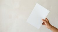 Male hand holding blank white paper sheet against neutral beige background, suitable for mockups, presentations, advertising campaigns, and design templates.