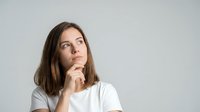 Thoughtful young woman with hand on chin looking up contemplatively against gray background, expressing curiosity and decision-making process.