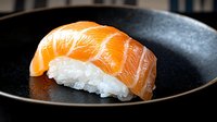 Fresh salmon nigiri sushi piece on black ceramic plate with glossy raw fish and perfectly pressed rice, dramatic lighting highlights texture and color.