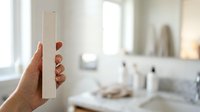 Woman holding clear glass test tube in modern bathroom with white fixtures and natural light from windows, suitable for healthcare, science, beauty, and wellness product marketing.