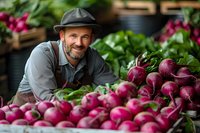 Mature Caucasian farmer in hat and apron smiling while displaying fresh organic red radishes at local farmers market stand surrounded by wooden crates and produce.