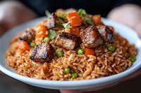 Grilled pork belly chunks served on aromatic fried rice with green peas, diced carrots and fresh herbs, close up view of Asian fusion cuisine on white plate.