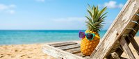 Cool pineapple with sunglasses relaxing on wooden beach chair by turquoise ocean water and sandy shore under blue sky with white clouds for summer vacation.