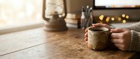 Cozy morning scene with hands holding steaming coffee mug on rustic wooden table near window with warm natural light and vintage lantern creating peaceful atmosphere.