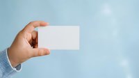Male hand holding blank white business card against light blue sky background with bokeh effect. Corporate identity mockup.