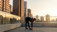 Athletic man stretching on urban rooftop terrace during golden hour sunset with city skyline backdrop for fitness and wellness lifestyle concepts.