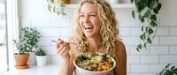 Happy blonde woman eating fresh healthy salad bowl in bright modern kitchen with plants and natural light for wellness lifestyle.