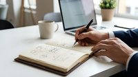 Professional businessman writing mathematical equations and formulas in notebook at modern office desk with laptop and coffee cup for business planning.