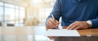 Middle-aged man with medium skin tone wearing dark blue shirt signing document at wooden desk in bright office environment with blurred background.
