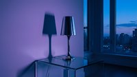 Two modern table lamps with cone-shaped shades on glass desk against purple neon-lit wall, city skyline visible through window at dusk, contemporary interior design.