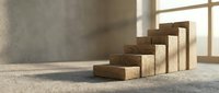 Wooden blocks arranged in ascending staircase pattern on concrete floor with sunlight from window, representing growth, progress, and business development concepts.