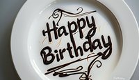 Happy Birthday message written in chocolate syrup on white ceramic plate with decorative swirls and flourishes, ready for celebration dessert presentation.