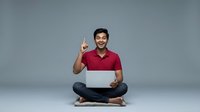 Young South Asian male in casual red polo shirt and dark jeans, sitting cross-legged while holding laptop, pointing upward with excited expression against neutral gray background.