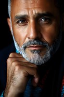 Close-up portrait of mature Middle Eastern man with gray beard in contemplative pose against dark background, dramatic lighting highlighting facial features.