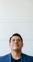 Middle-aged man of Latin American descent wearing clear-framed glasses and navy blazer against neutral white wall background. Professional headshot suitable for corporate, business, and portrait use.