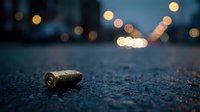 Single spent bullet casing lying on dark asphalt street at night with blurred city lights creating atmospheric bokeh background for crime scene concepts.