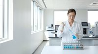 Young female scientist in white lab coat conducting research with test tubes in modern laboratory setting with equipment and large windows.