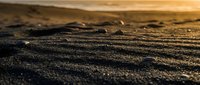 Golden sunset light illuminates textured sand patterns on beach with gentle waves in background creating warm atmospheric coastal scene for travel content.