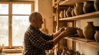 Senior male potter crafting ceramic vessel in traditional workshop studio with clay pots on wooden shelves by window creating handmade pottery art.