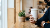 Close-up of a woman's hand holding a blank white takeaway coffee cup in a modern cafe interior with potted succulent plant and blurred background.
