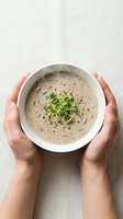 Overhead view of hands holding white bowl with creamy mushroom soup garnished with fresh microgreens and black pepper, ready for serving or food photography.