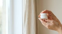 Adult hands holding blank white cosmetic jar against window with natural light and neutral beige curtains, suitable for skincare product packaging mockups and beauty branding.