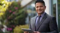 Confident mature businessman in gray suit holding digital tablet outdoors with blurred garden background, representing corporate success and leadership.