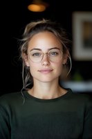 Young Caucasian woman wearing round glasses and green sweater smiles warmly at camera. Natural makeup, loose updo hairstyle with wispy strands, soft lighting creates intimate portrait.