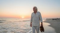 Senior man with gray hair and light skin tone, wearing casual white linen shirt, walking barefoot on sandy beach at sunset holding shoes. Peaceful retirement lifestyle and coastal leisure.