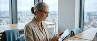 Mature woman with gray hair and glasses wearing beige blazer holding tablet in modern high-rise office with city skyline view, business professional setting.