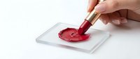 Caucasian female hand applying red lipstick on clear acrylic swatch, smearing bold crimson pigment for cosmetic testing on white background.