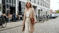 Confident blonde businesswoman in beige coat walking on European city street with bicycles and outdoor cafe. Professional urban lifestyle concept.