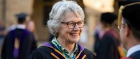 Smiling senior Caucasian woman in graduation cap and gown with glasses celebrating academic achievement at university ceremony with proud expression.