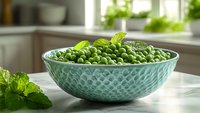 Fresh green peas in turquoise ceramic bowl with mint leaves on white kitchen counter near window, natural daylight creates appetizing food photography setup.