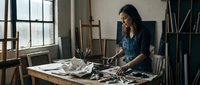 Young female artist with long dark hair working on paintings in bright studio workspace with easel, brushes and art supplies on wooden table.