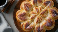 Freshly baked flower-shaped sweet bread roll with golden brown crust and poppy seeds on wooden plate, served with coffee and cinnamon sticks.