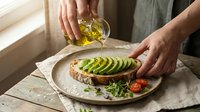 Hands drizzling olive oil over sliced avocado toast on rustic plate. Healthy breakfast preparation with fresh herbs and cherry tomatoes on wooden table.