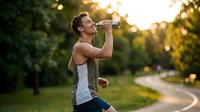 Caucasian male athlete drinking water from bottle during outdoor running workout in park at sunset, refreshing hydration break.
