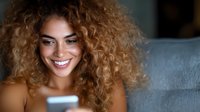 Young mixed race woman with curly hair smiling while looking at mobile phone screen in dark room illuminated by blue light, expressing joy and engagement.