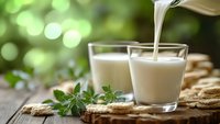Fresh milk being poured into glass on rustic wooden table with green herbs and natural bokeh background for healthy breakfast and organic dairy products.