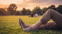 Man relaxing on green grass in a park at sunset, legs in casual khaki pants and suede loafers, golden hour bokeh background.