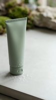 Pale green cosmetic tube with ribbed cap displayed on white stone surface with water droplets, blurred natural background with moss and rocks.