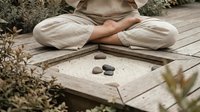 Woman in beige linen clothing sitting cross-legged on wooden deck practicing meditation with zen stones arranged on sand tray surrounded by dried plants.