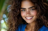Young mixed race woman with curly brown hair and bright smile outdoors. Natural freckles, white teeth, and blue top against blurred green background.