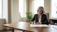 Mature businesswoman with gray hair writing documents at wooden desk in bright modern office with large windows and plants.