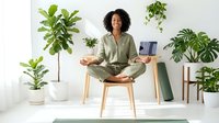 Woman with curly dark hair and medium skin tone meditating in lotus position on wooden chair surrounded by potted plants and laptop, promoting wellness and mindfulness in home office environment.