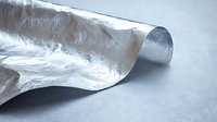 Curled sheet of silver aluminum foil with textured metallic surface reflecting light against neutral gray background, creating dynamic waves and shadows.
