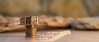 Miniature wooden house model on stack of coins with rising percentage arrows showing real estate investment growth and property market trends.