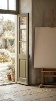 Artist studio interior with open wooden door, blank canvas on easel, and potted plants by window with natural light.